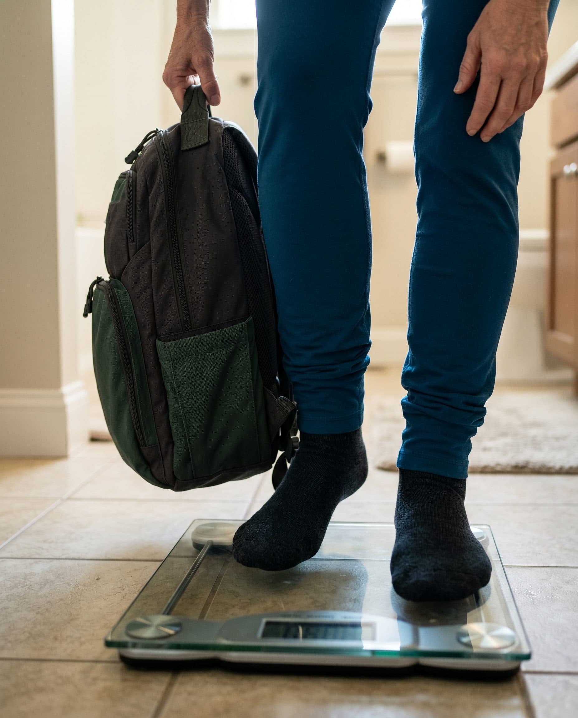 A woman in teal leggings and socks steps onto a bathroom scale, holding a backpack by its handle.